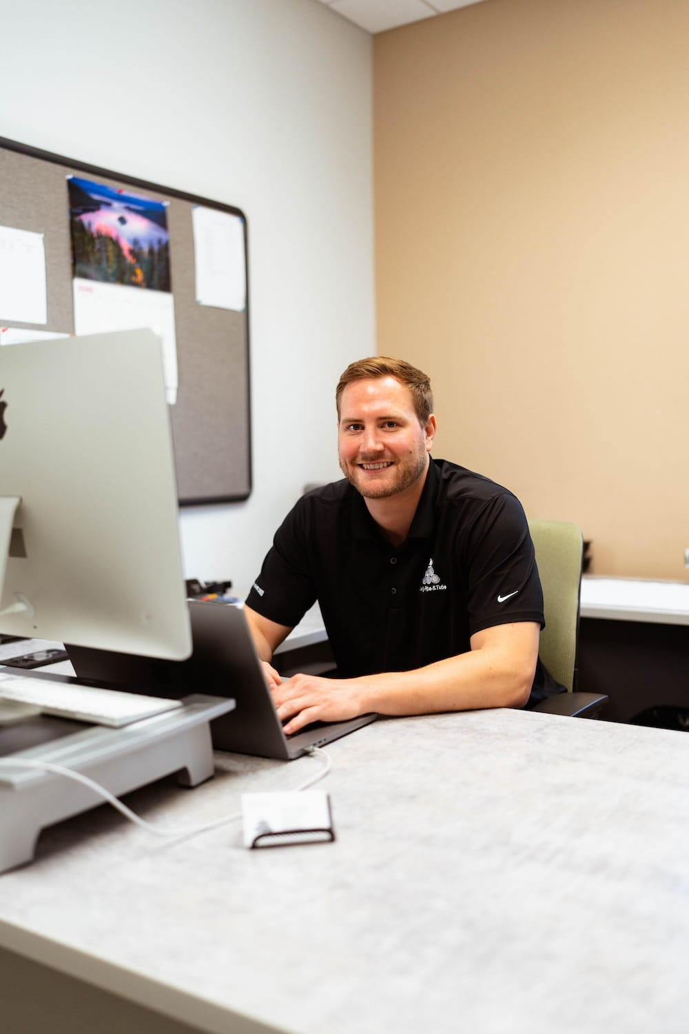 Smiling pipe and tube industry sales representative working at a desk with a laptop and iMac computer in a professional office environment