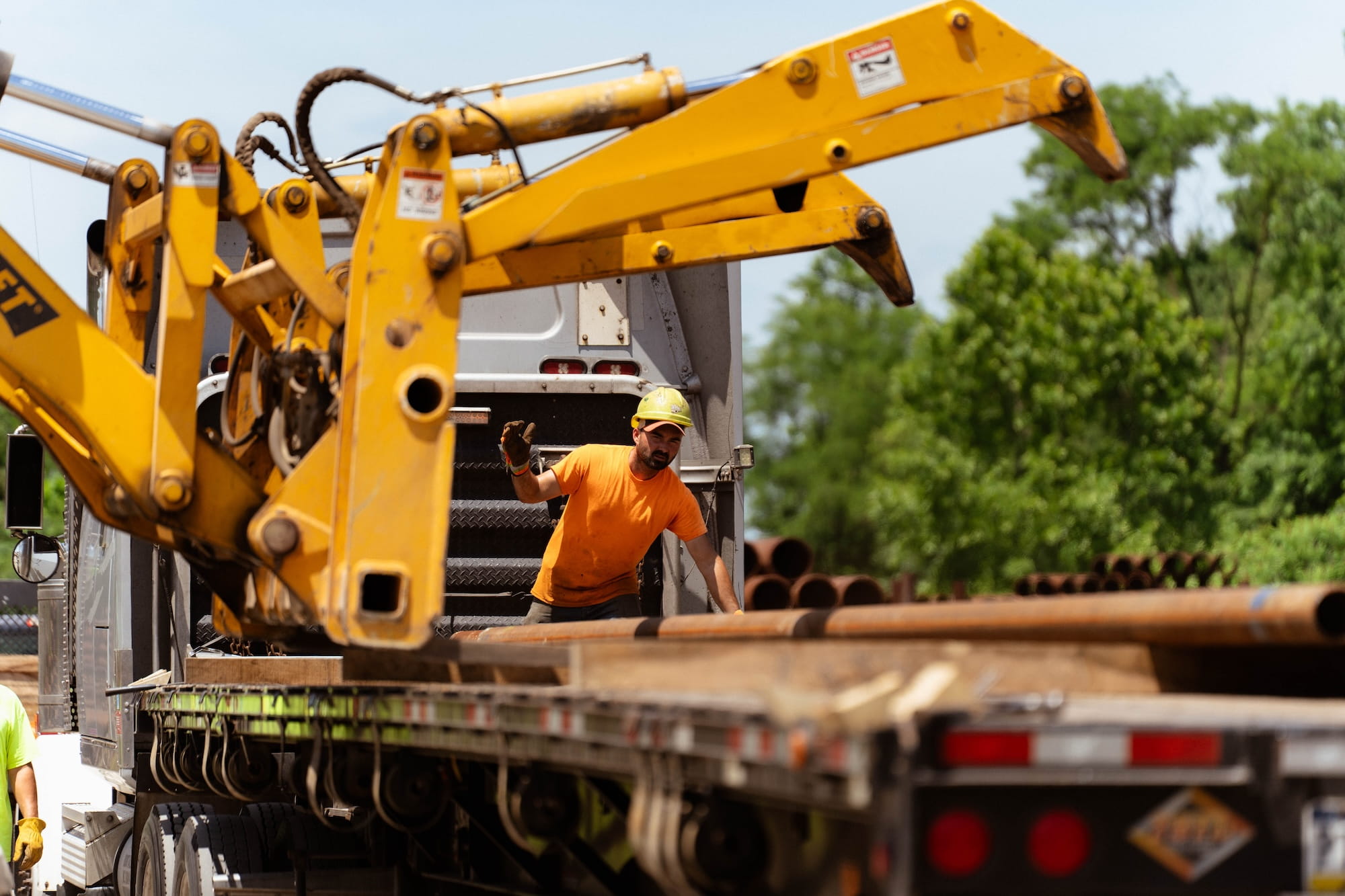 Pipe yard worker in a yellow hard hat and orange hi-vis shirt directing a pipe handler crane while loading steel pipe onto a flatbed truck for delivery