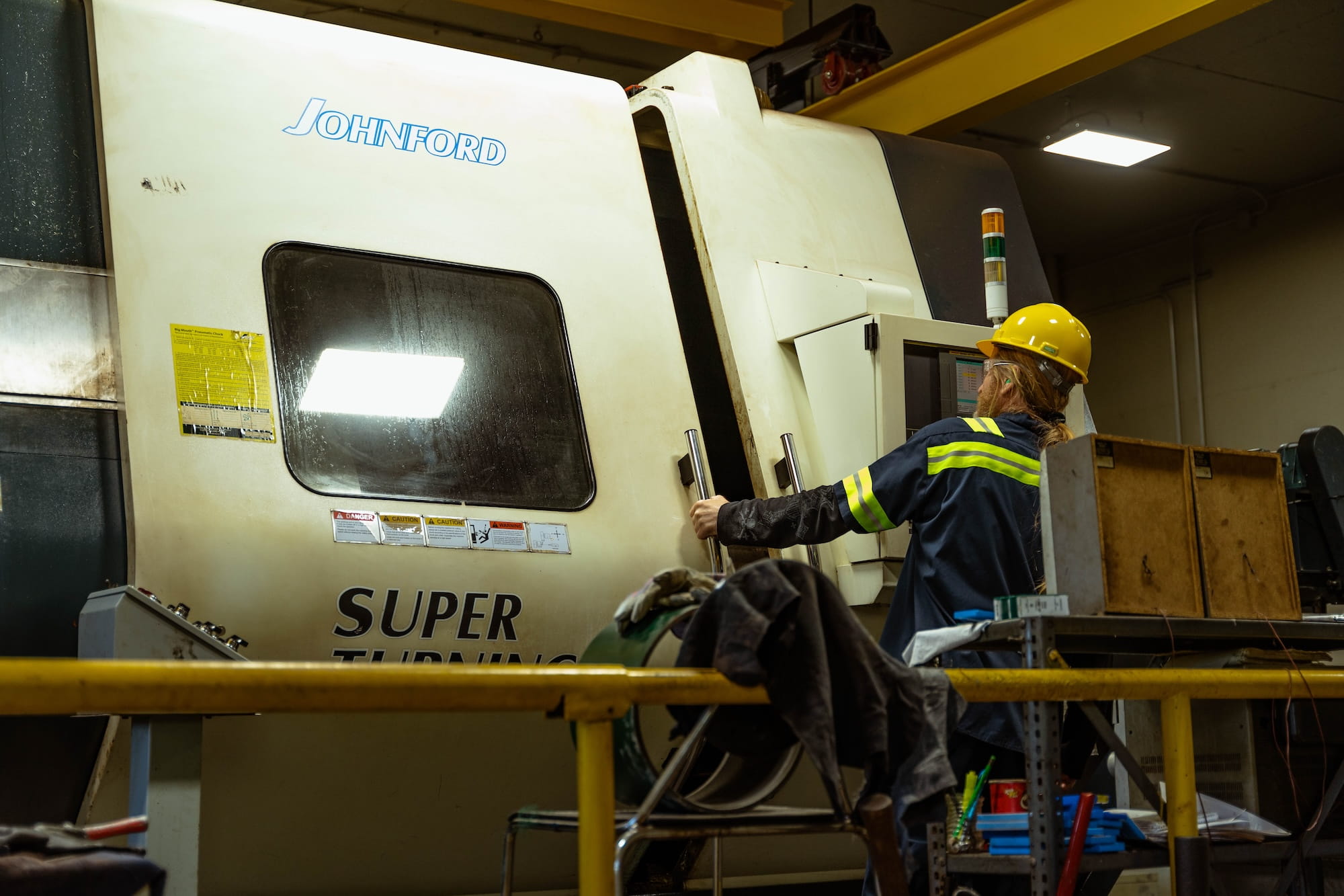machinist in a yellow hard hat and hi-vis safety gear operating a Johnford CNC super turning lathe machine in an industrial pipe threading facility
