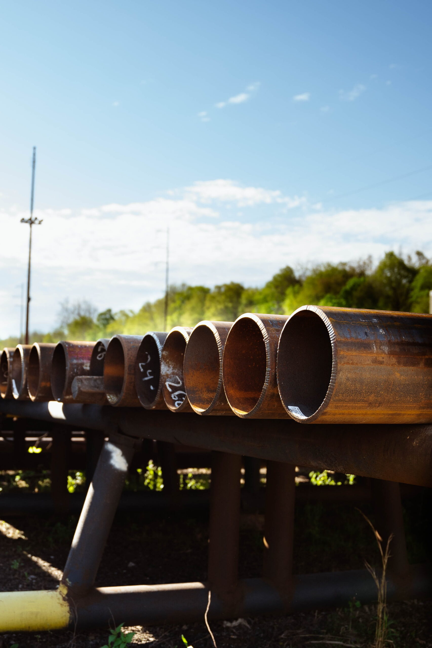Row of used carbon steel pipes with beveled ends stored on outdoor pipe racks at an industrial pipe yard with trees and blue sky in the background