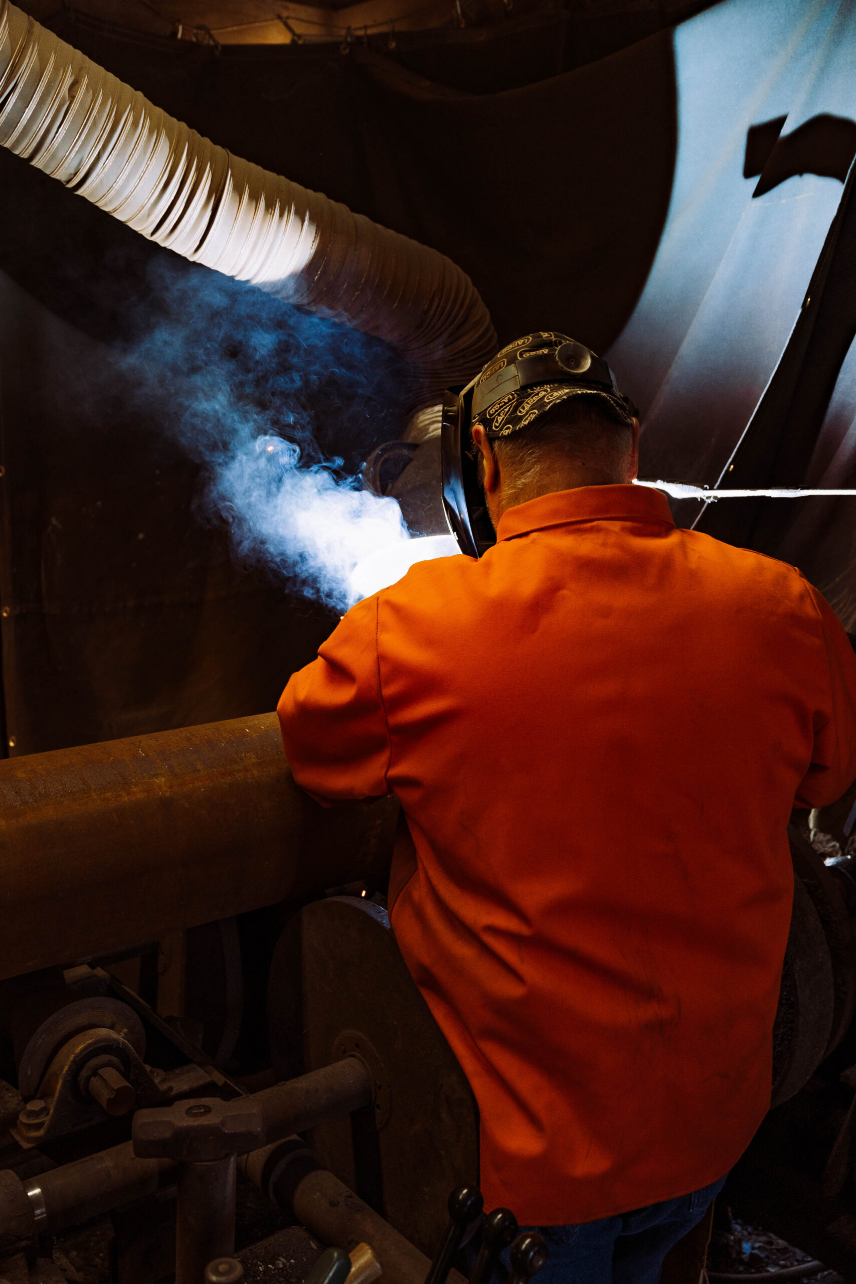 Pipe welder in an orange uniform and welding helmet actively welding steel pipe inside an industrial pipe fabrication shop with welding smoke and bright arc light visible