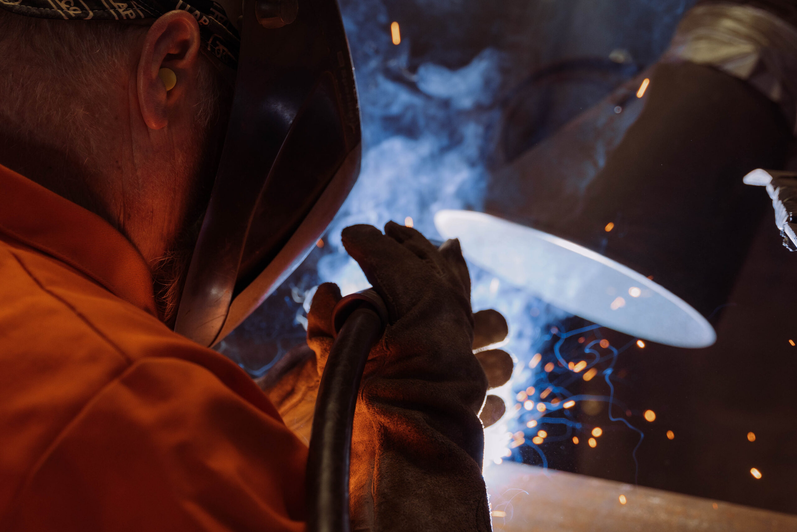 Extreme close-up of a pipe welder with leather gloves and ear protection welding steel pipe with bright arc light, sparks, and smoke in an industrial pipe fabrication shop