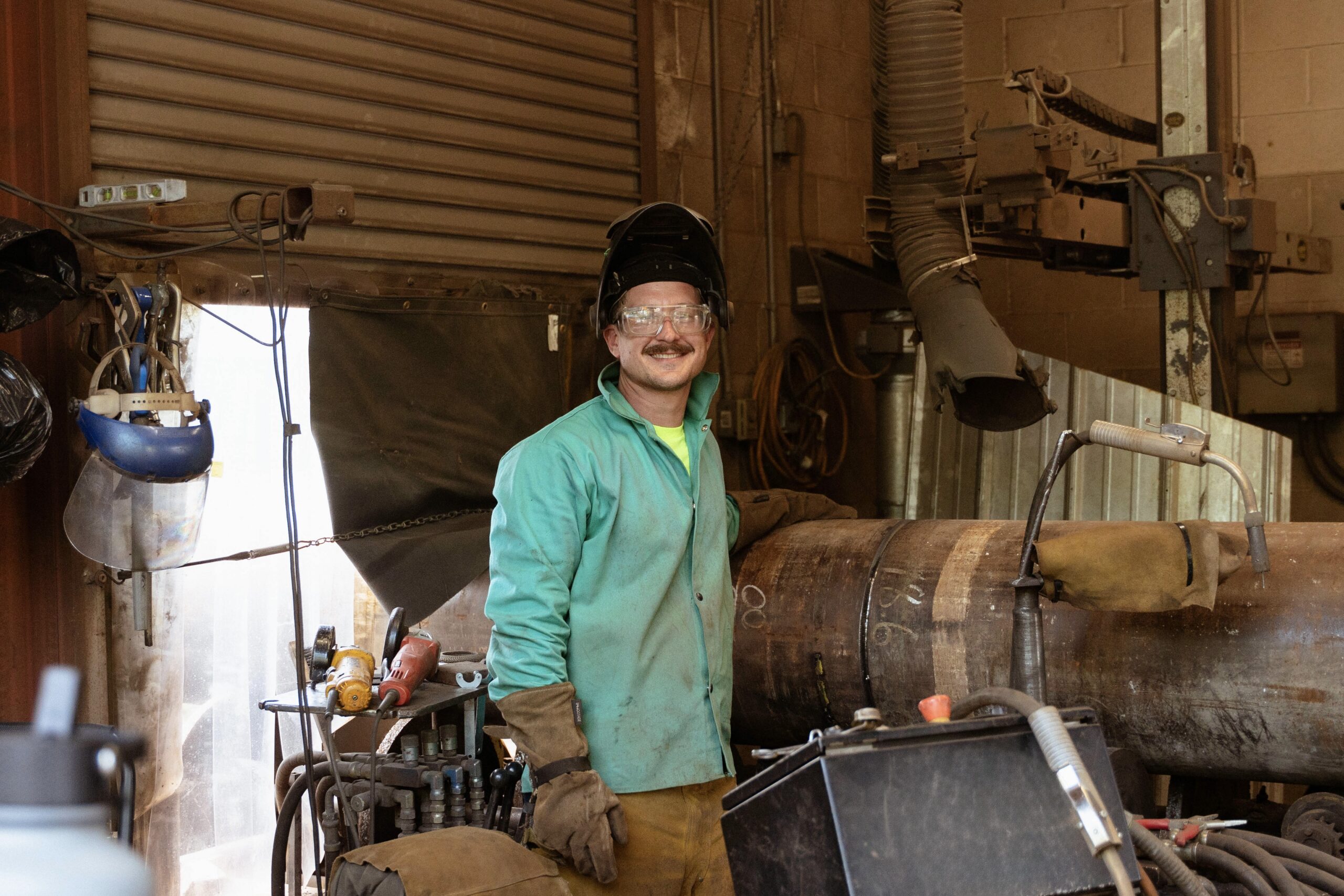 Pipexperts welder smiling in a green welding jacket, safety glasses, and welding helmet standing next to industrial steel pipe in an indoor pipe fabrication shop
